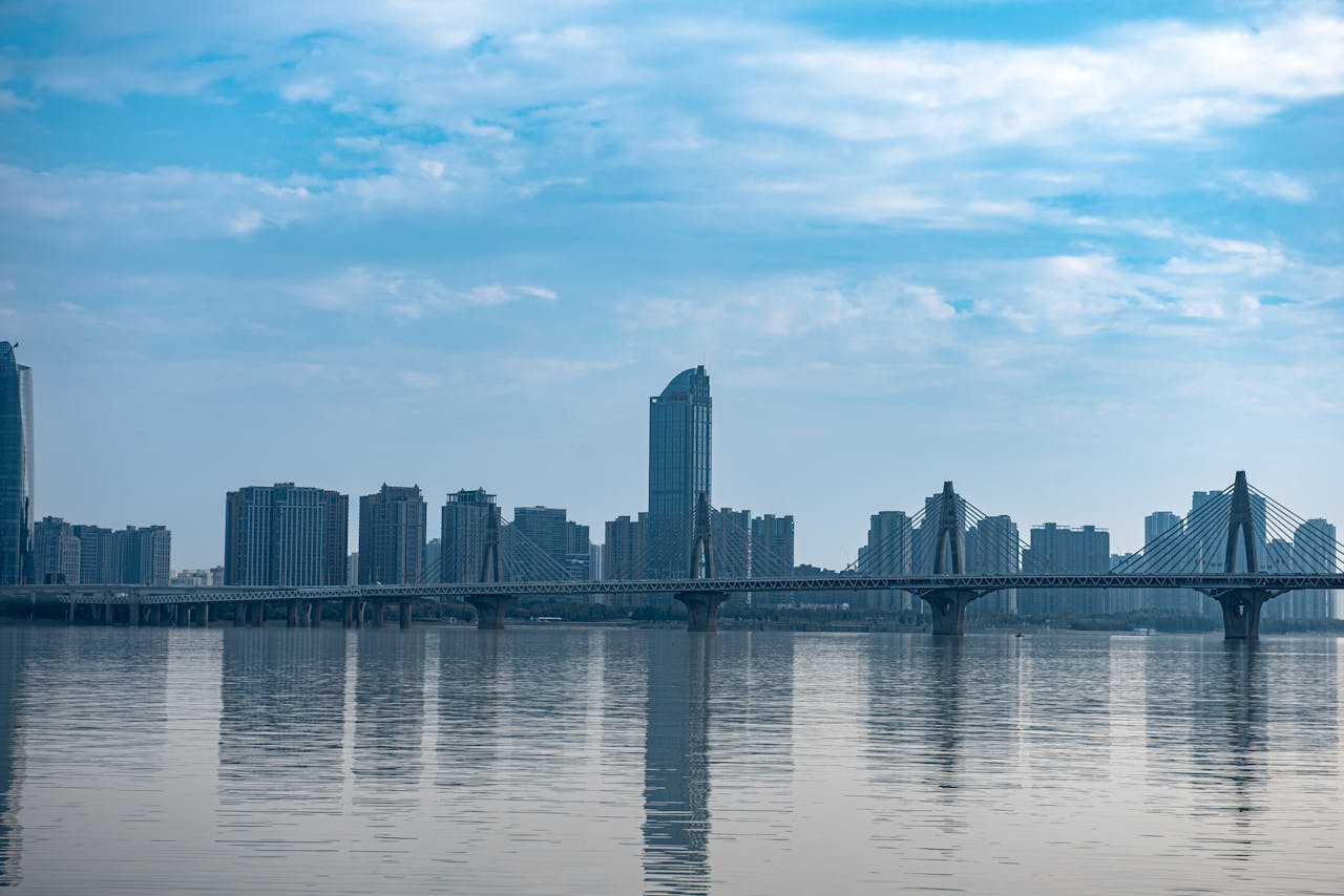 A panoramic view of a modern city skyline featuring a bridge over calm water under a blue sky.